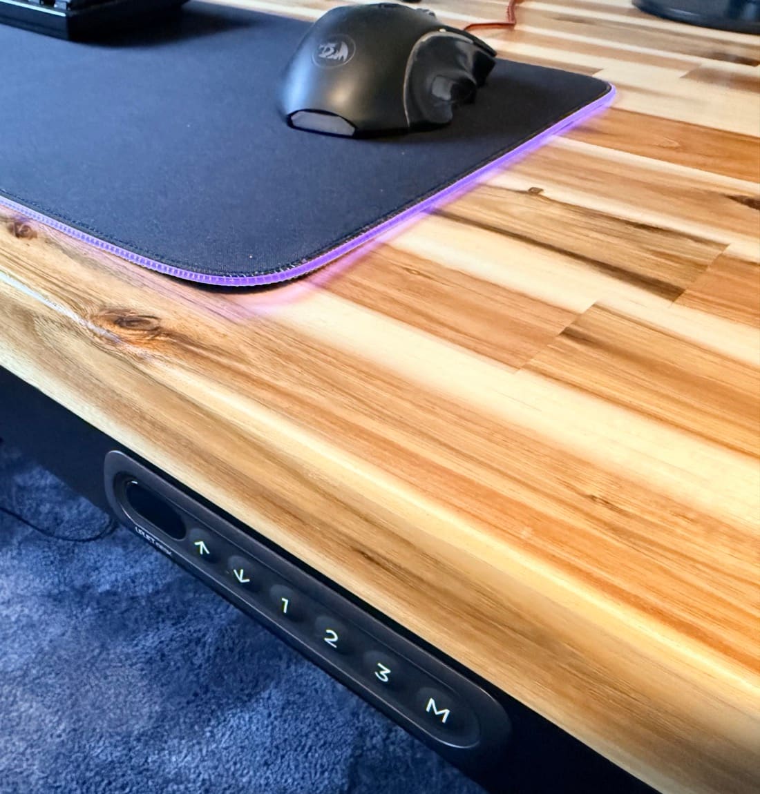 UPLIFT Parsons Standing Desk in daily use, showing the flush keypad embedded in the front apron, Acacia butcher block desktop, and lived-in setup with desk mat and mouse