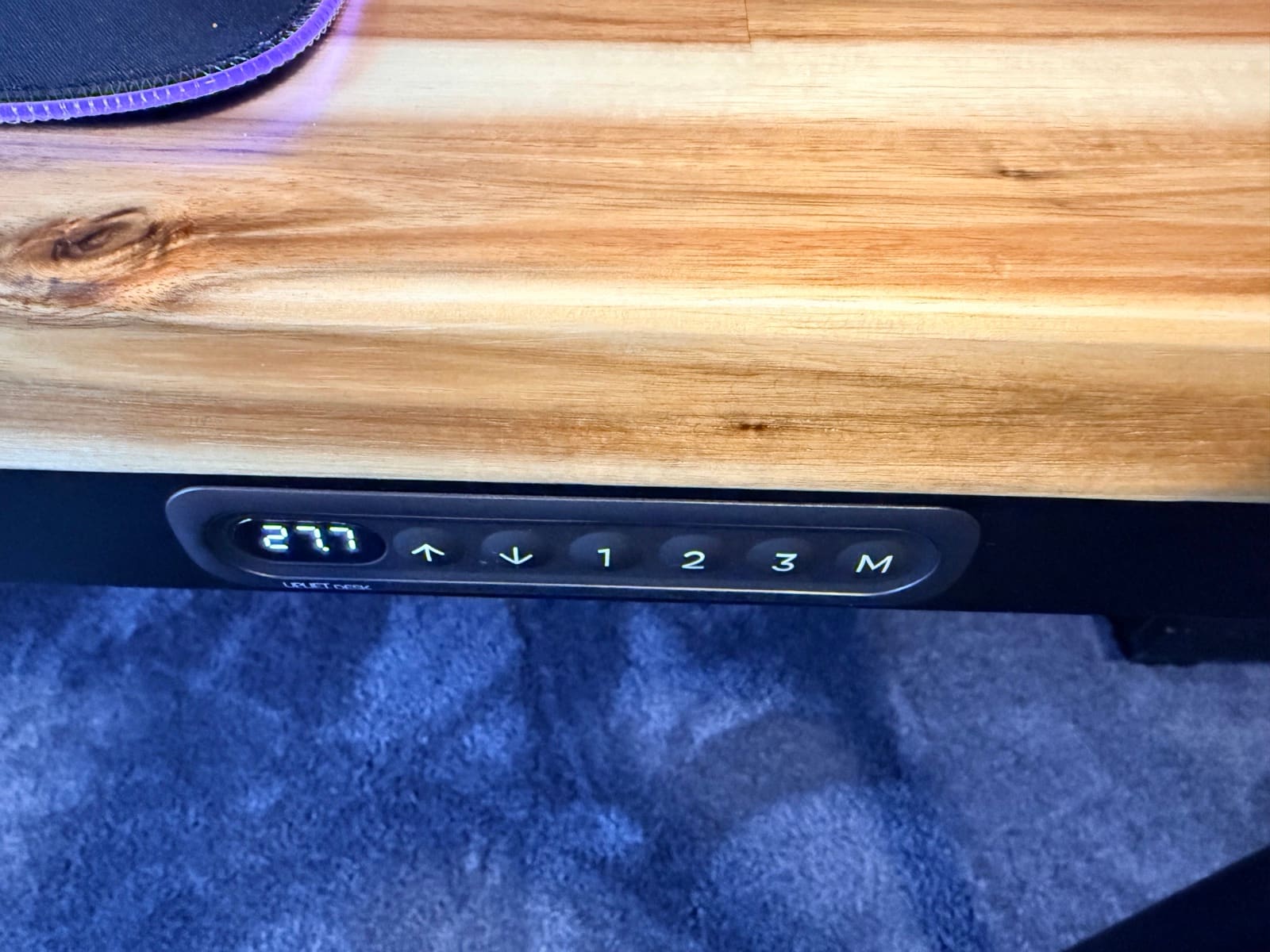 UPLIFT Parsons Standing Desk keypad close-up showing the height display reading 27.7 inches, with up and down arrows, three memory presets, and an M button, all integrated flush into the front apron of the Acacia butcher block desktop