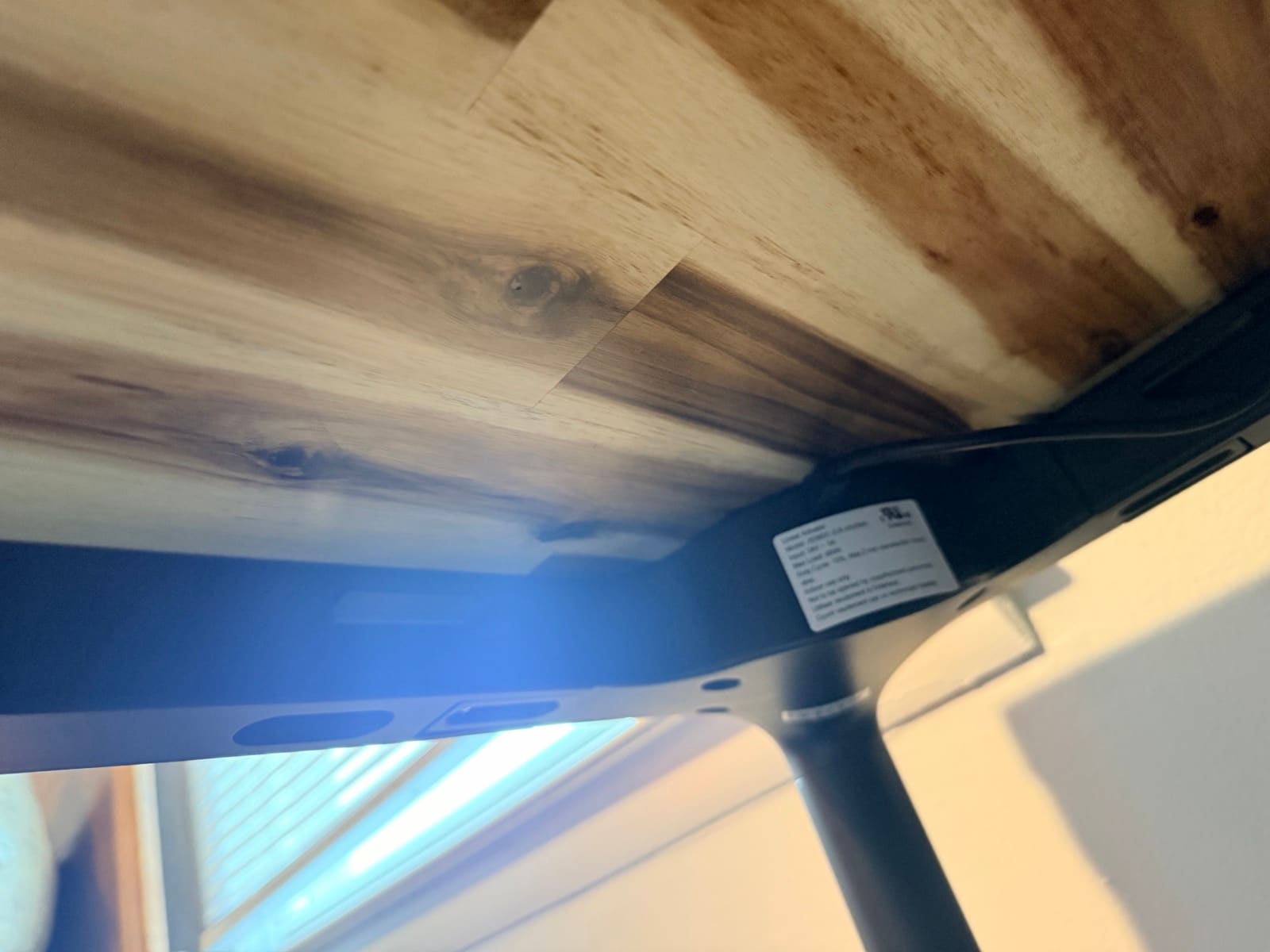 Under-desk view of the UPLIFT Parsons Standing Desk showing the control box, UL safety label, cable channel, and tapered leg meeting the underside of the Acacia butcher block desktop