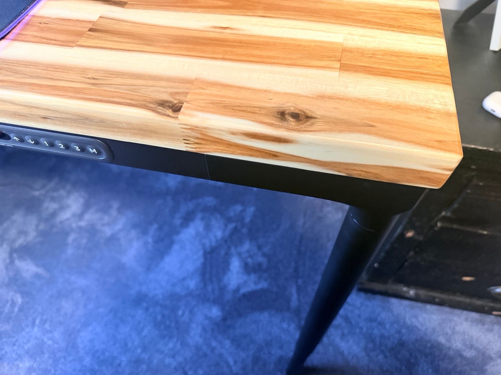 UPLIFT Parsons Standing Desk shown at a low angle in a real room, with the tapered leg meeting the Acacia butcher block desktop and a piece of furniture visible in the background, showing how the desk integrates into a room with other furniture