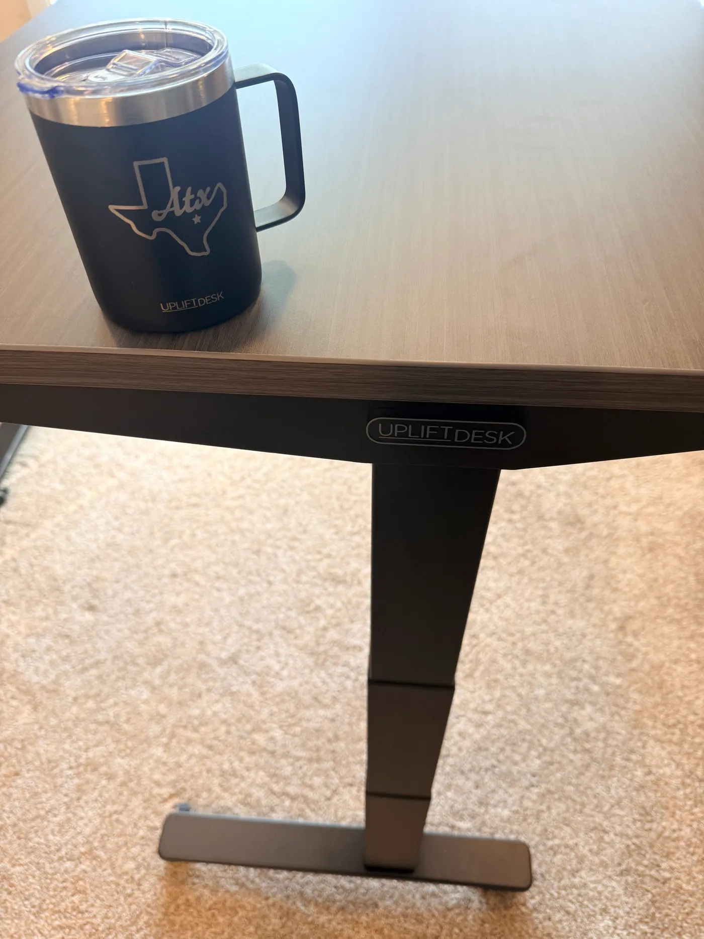 The branded UPLIFTDESK logo plate mounted on the V3 frame leg, alongside the Texas-shaped insulated UPLIFT mug from the accessories box